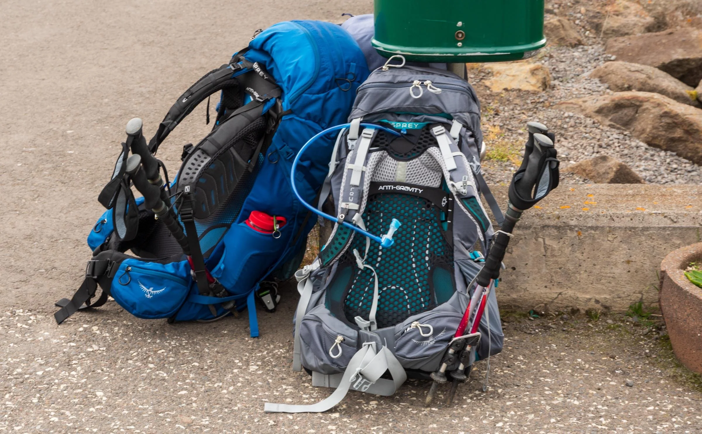 A collection of colorful backpacks and hiking gear placed on the ground outdoors near rocks, with a green trash bin in the background.