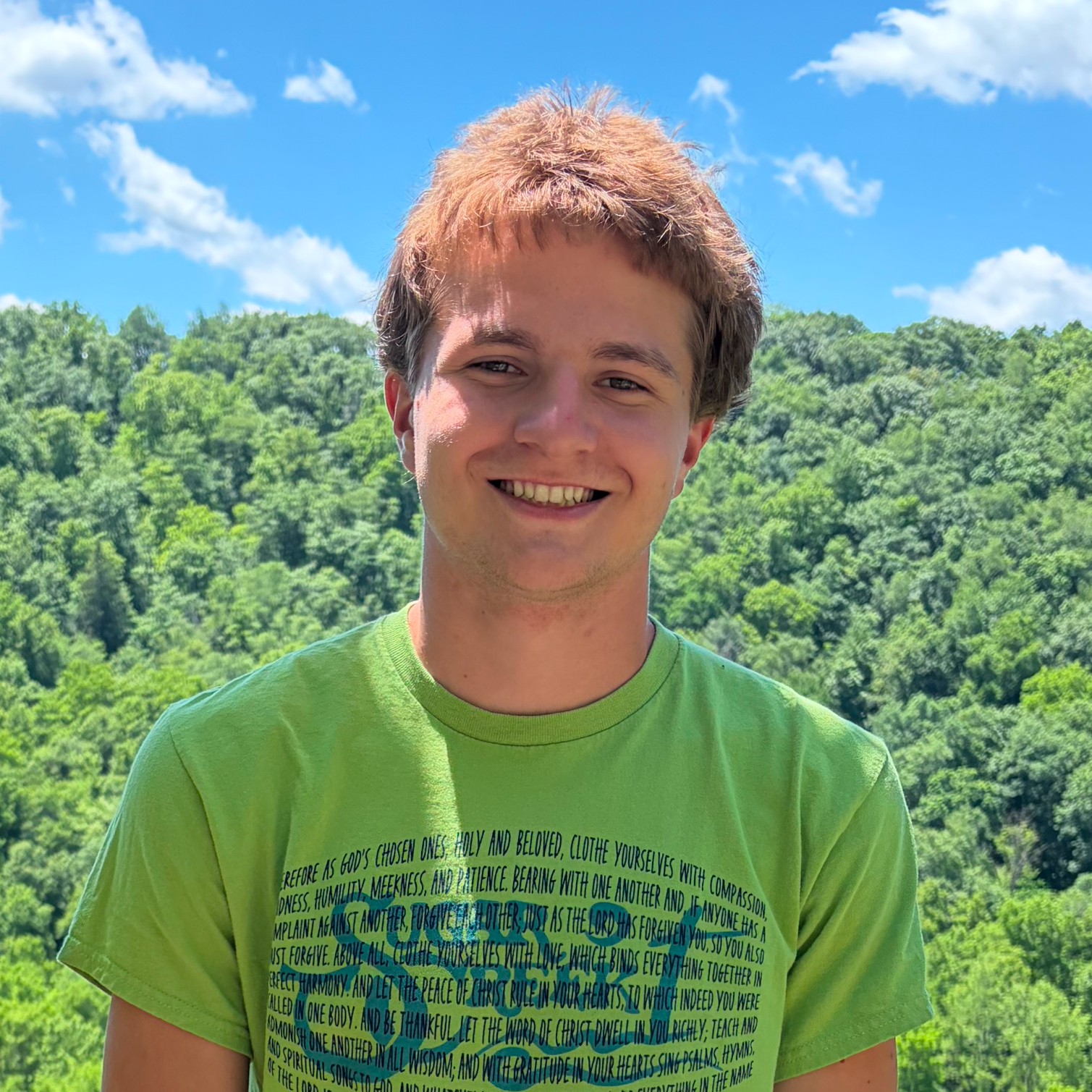Young man smiling outdoors with a green background, wearing a bright green T-shirt with printed text on it and a scenic blue sky with clouds behind him.