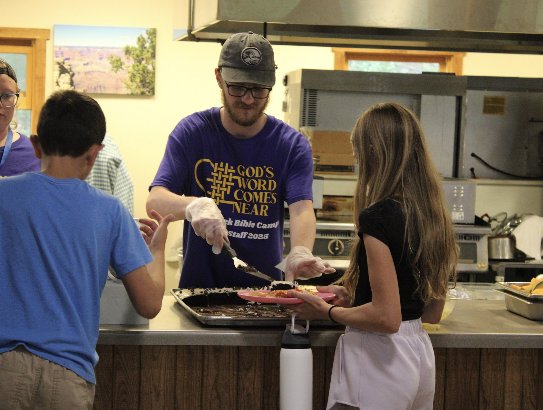 A group of people serving and receiving food in a kitchen. There are boys and girls and an adult man in a purple T-shirt, which says 'God's Word Comes Near' and 'Biblical Camp Staff 2025.' One girl in a black top and pink shorts is handing a plate of cake to an adult man. The scene is indoors with kitchen equipment and colorful pictures on the walls.