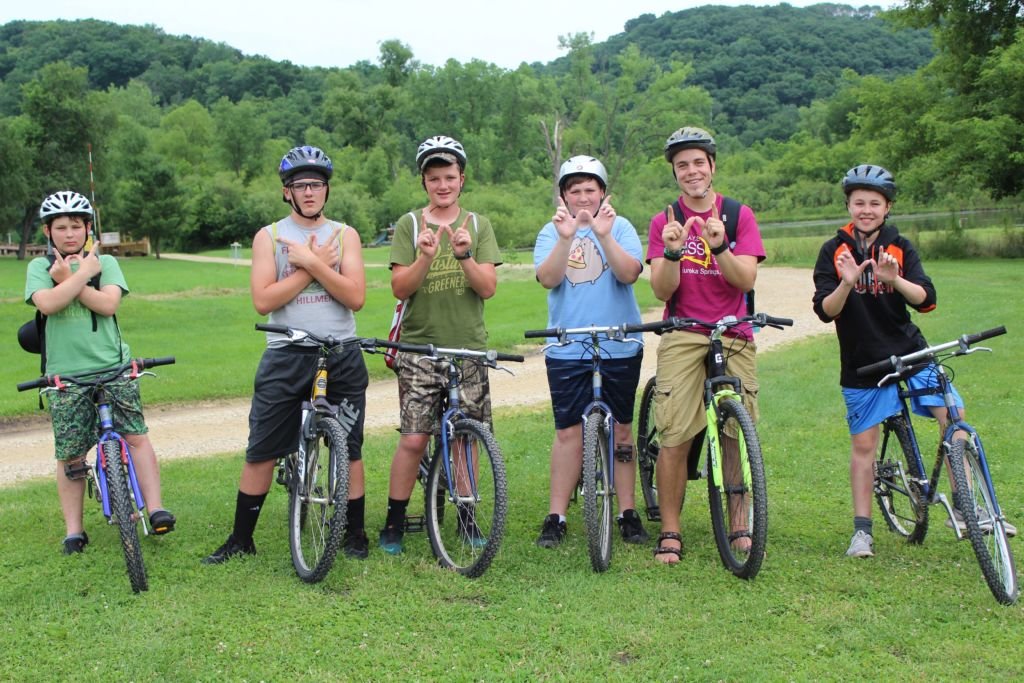 Six children wearing helmets and casual clothing standing with bikes on a grassy field with trees and hills in the background.