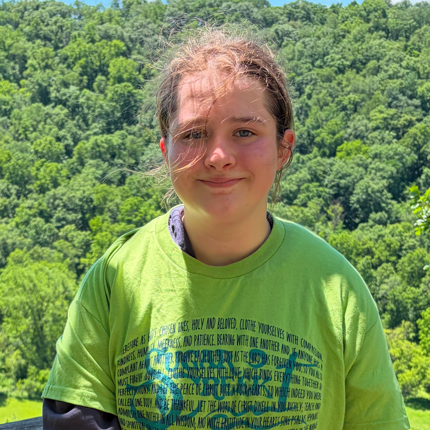 A young person with light skin and curly hair, wearing a bright green T-shirt with text on it, standing outdoors in front of lush green trees under bright sunlight.