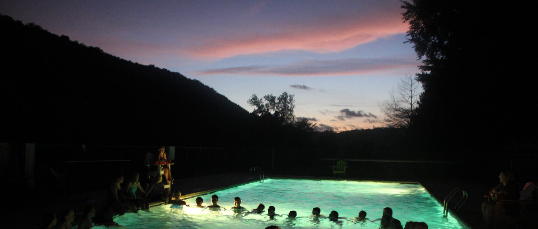 People swimming in an outdoor pool during sunset with trees and hills in the background.