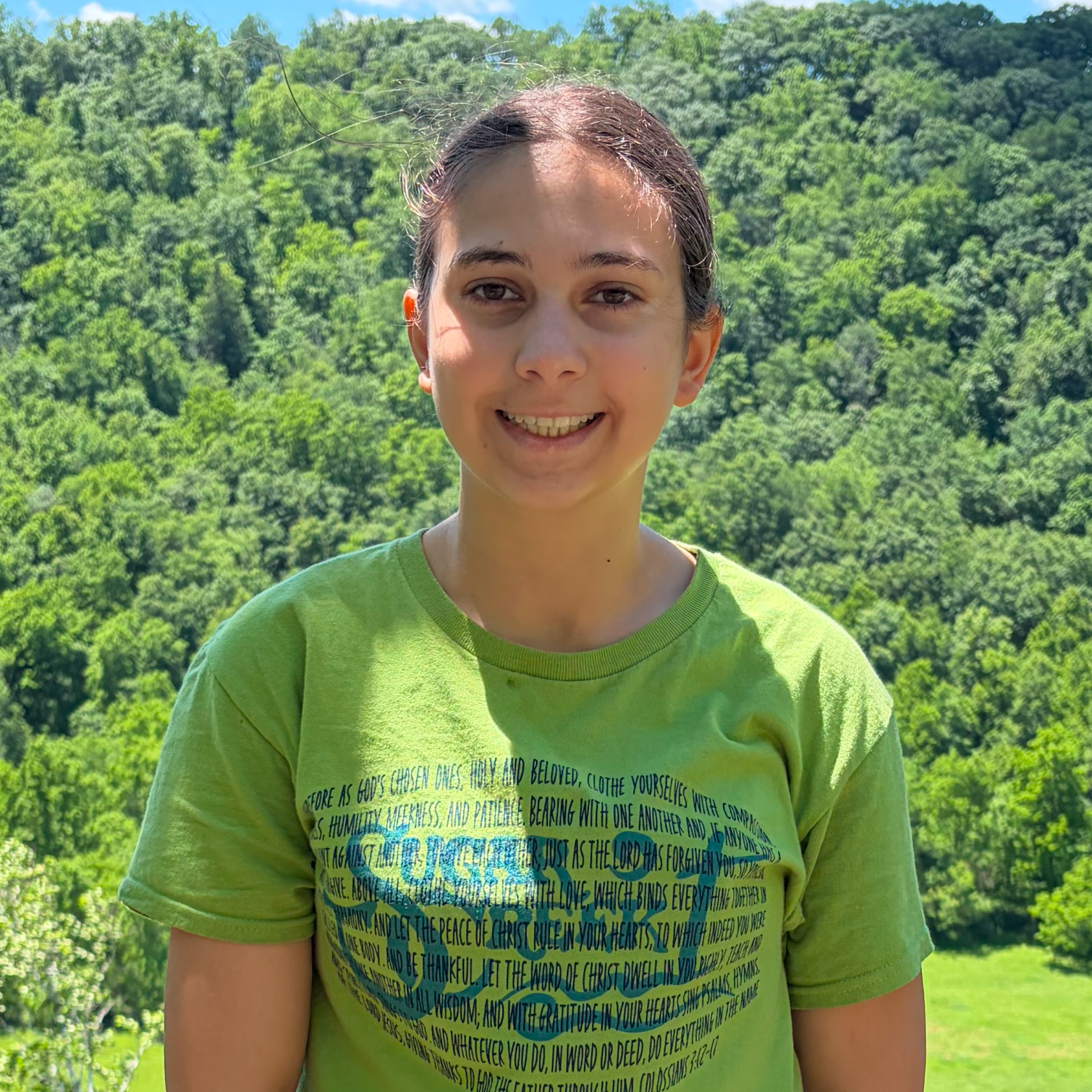 A smiling girl with dark hair tied back, wearing a light green T-shirt, in front of a lush green forest background on a sunny day.