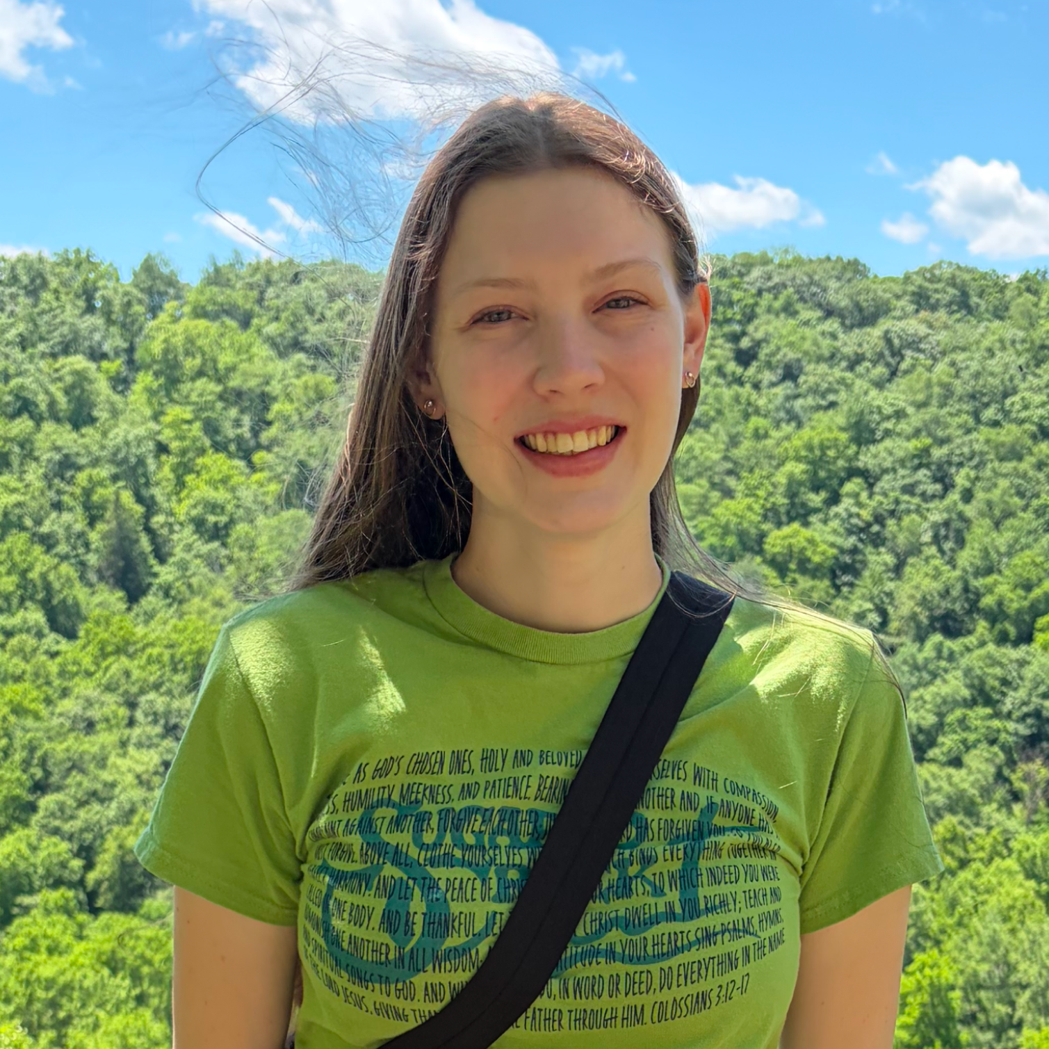 A young woman with long dark hair, smiling, wearing a green T-shirt with text, and a black crossbody bag, standing outdoors in a green landscape with trees under a bright blue sky.