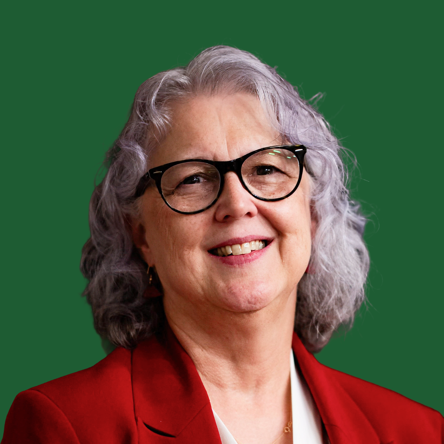 A close-up portrait of a smiling woman with gray, curly hair, wearing glasses, a red blazer, and a white shirt, against a solid green background.