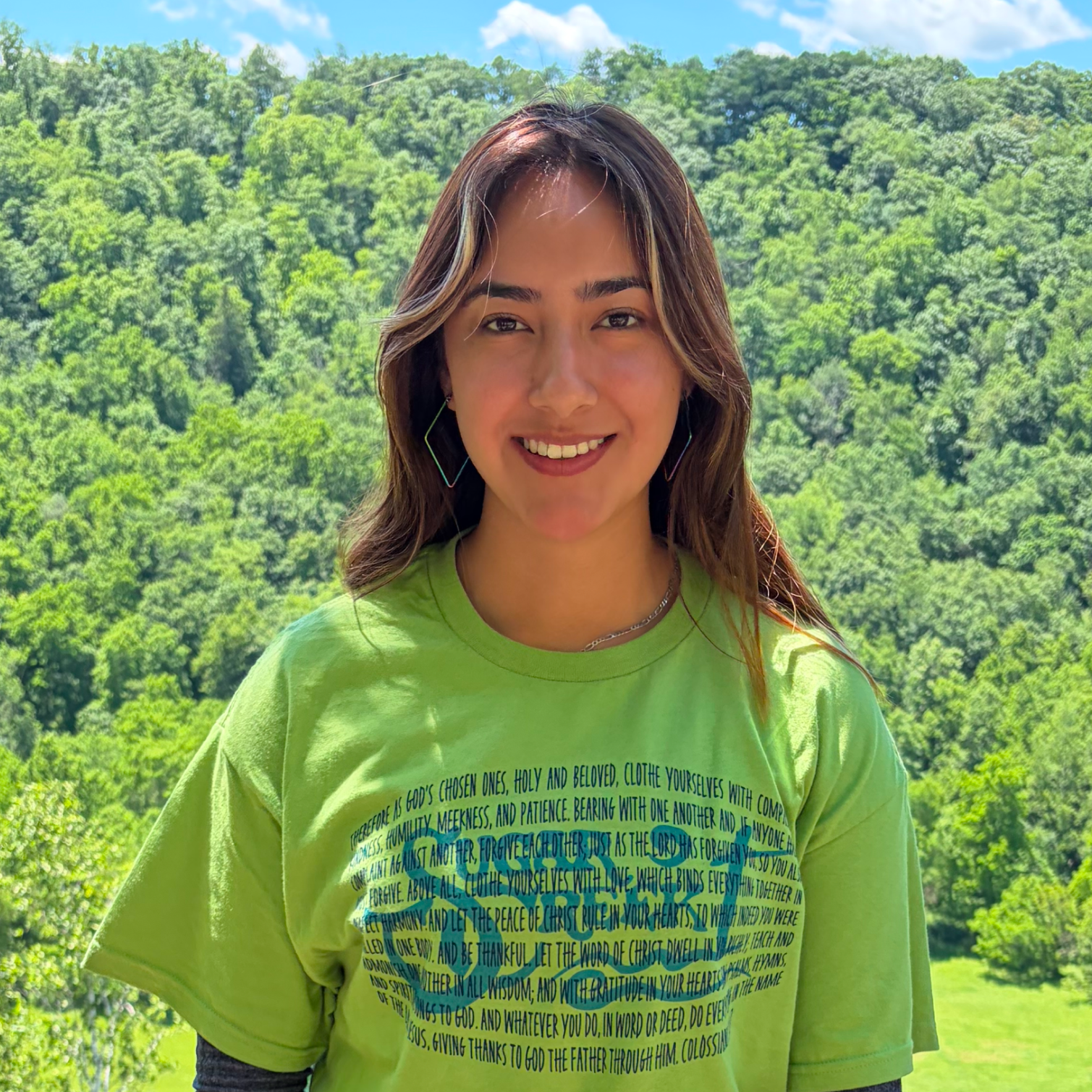 A young woman smiling outdoors with a green forested background, wearing a bright green t-shirt with a Bible verse written on it and large, colorful earrings.