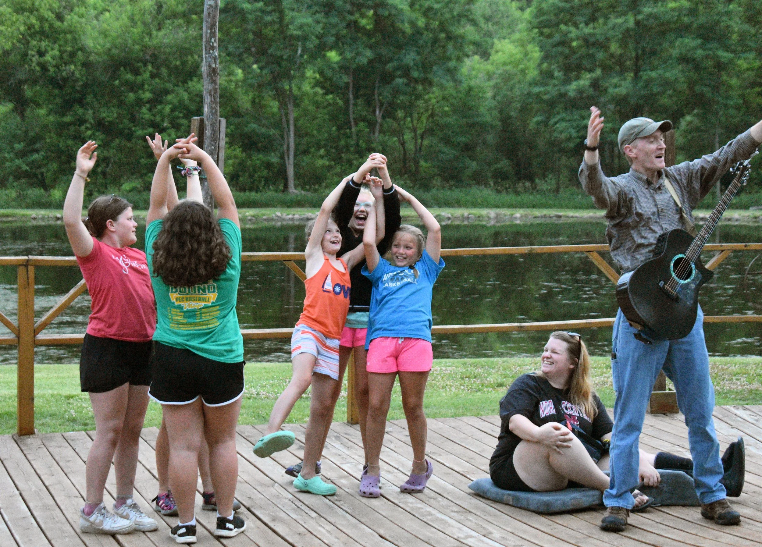 A group of children and an adult happily dancing and singing outdoors on a wooden deck near a body of water, surrounded by green trees.