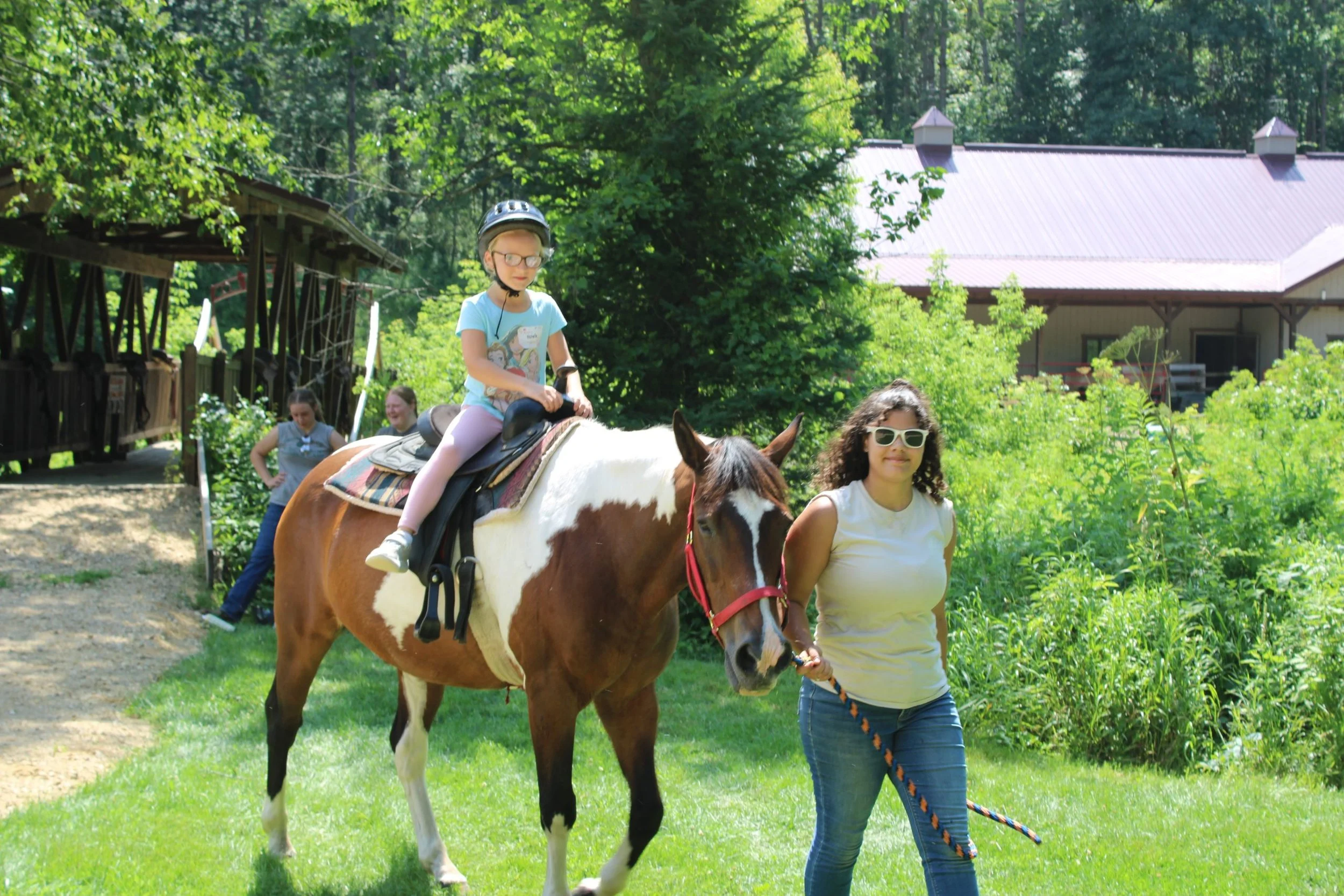 A young girl with glasses and a helmet riding a brown and white horse at an outdoor park, with two women walking alongside and smiling, surrounded by greenery and trees.