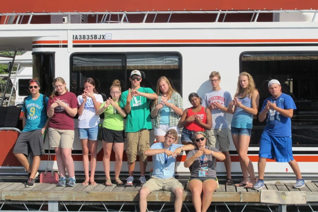 A group of young people posing in front of a large white boat with black and orange stripes, on a dock with a wooden platform.