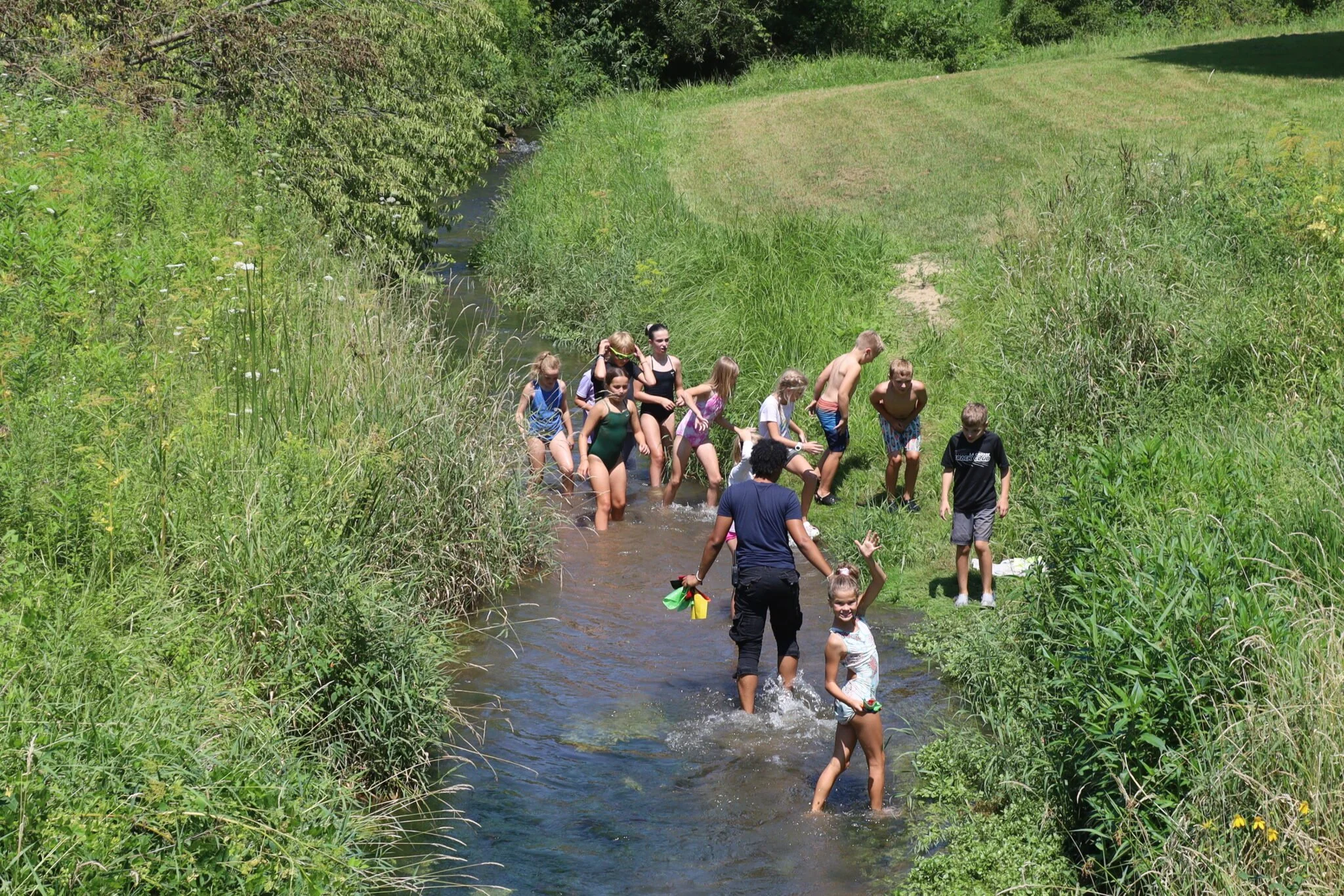 A group of children and teenagers wading through a shallow stream in a lush, green outdoor setting on a sunny day.