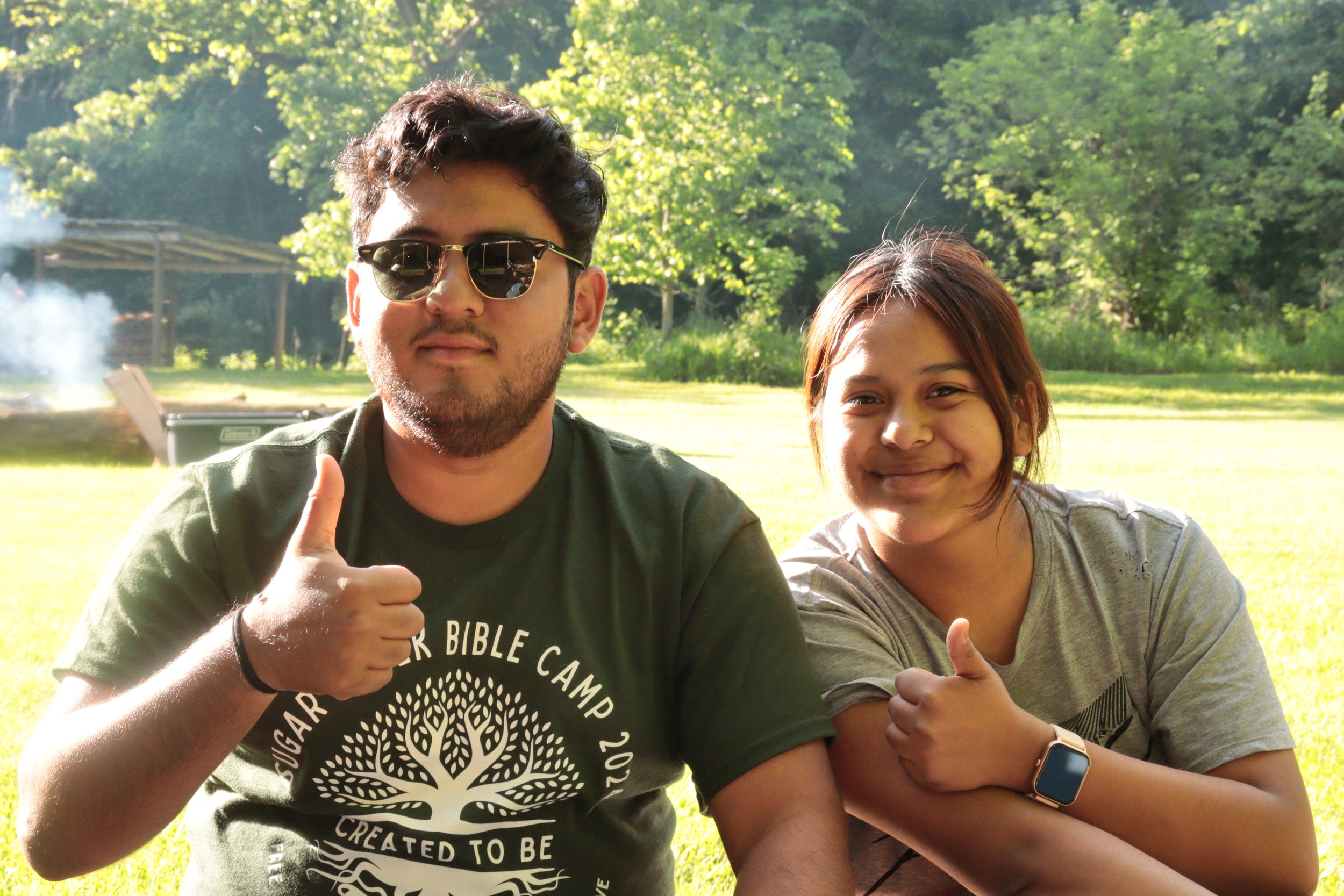 Two people, a man and a woman, sitting outdoors in a park with green trees in the background. The man is wearing sunglasses and a dark T-shirt, giving a thumbs-up. The woman is smiling and also giving a thumbs-up, wearing a smartwatch on her left wrist.