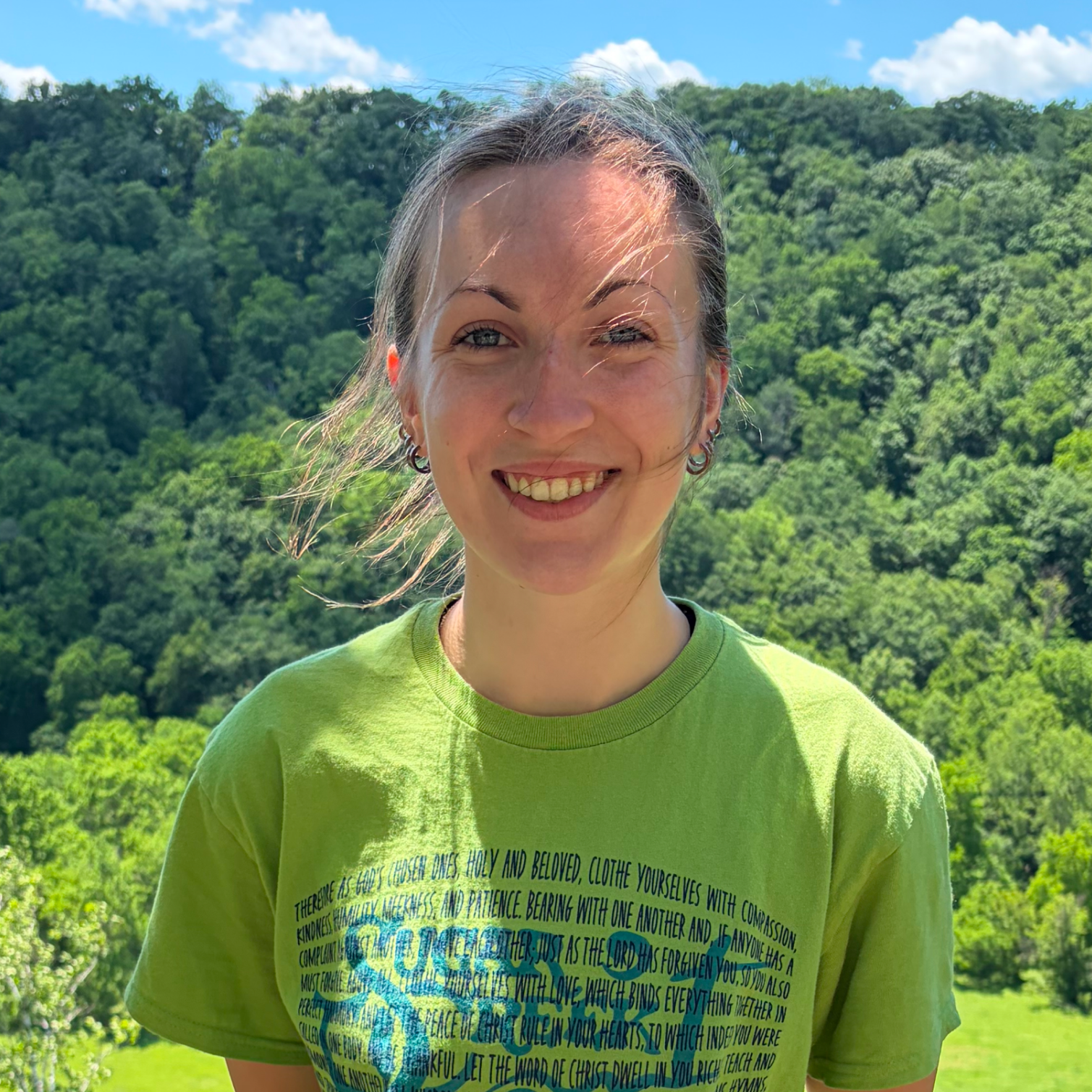 A smiling woman with windblown hair stands outdoors against a background of green trees and a blue sky with white clouds. She is wearing a bright green T-shirt with a Bible verse printed on it.