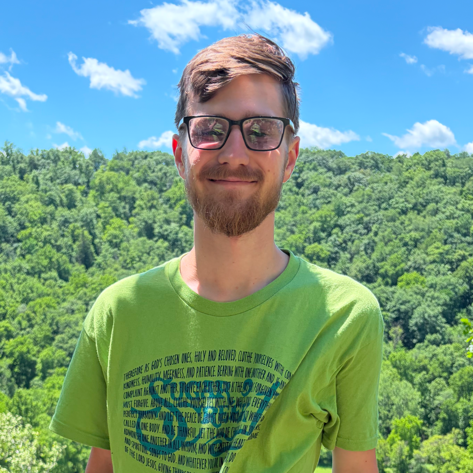 A young man with brown hair, beard, and glasses is smiling and standing outdoors against a backdrop of green trees and a partly cloudy blue sky, wearing a bright green T-shirt with text printed on it.