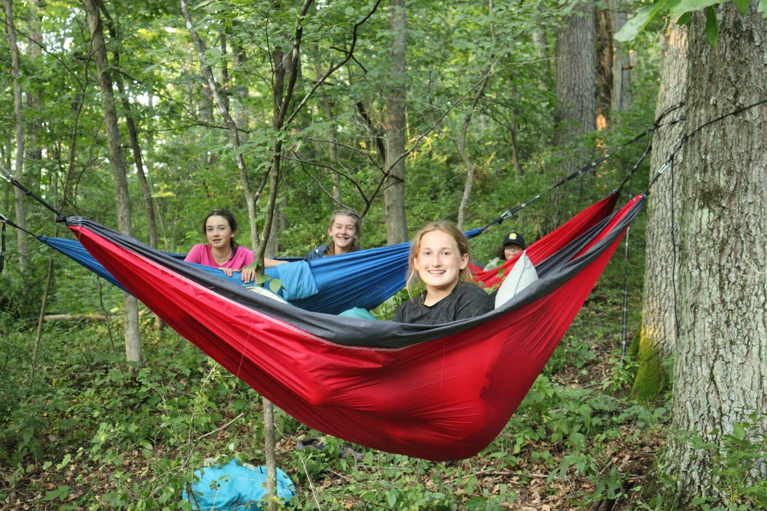 Four young girls smiling and sitting in a large red, blue, and black hammock tied between two trees in a wooded area.
