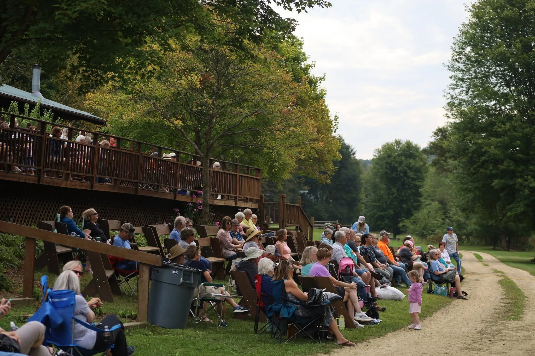 People sitting on tiered wooden benches outdoors, watching an event, with trees and a cloudy sky in the background.