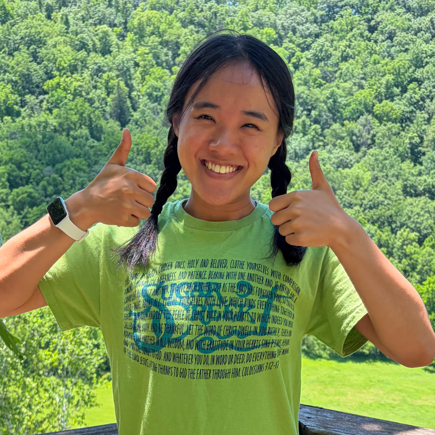 A young woman with long black braided hair giving two thumbs up, smiling, wearing a bright green T-shirt with a Bible verse, on a wooden deck with a lush green forest in the background.