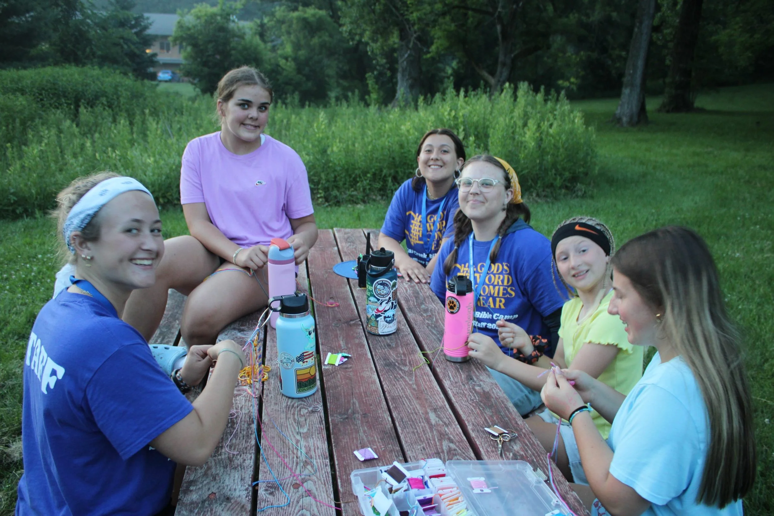 A group of young girls gathered around a wooden picnic table outdoors, engaged in a craft activity. They are smiling, with some holding craft supplies. The setting is a grassy area with trees in the background, suggesting a park or camp environment.