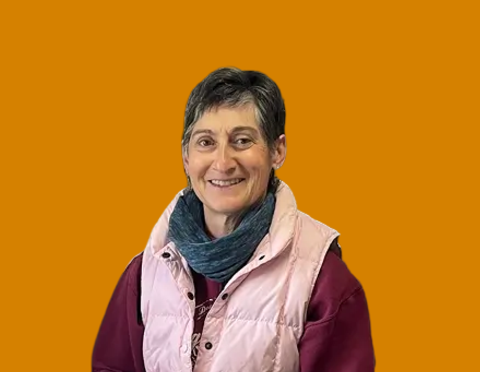 A woman with short dark hair smiling, wearing a pink vest over a maroon shirt and a blue scarf, against a solid orange background.