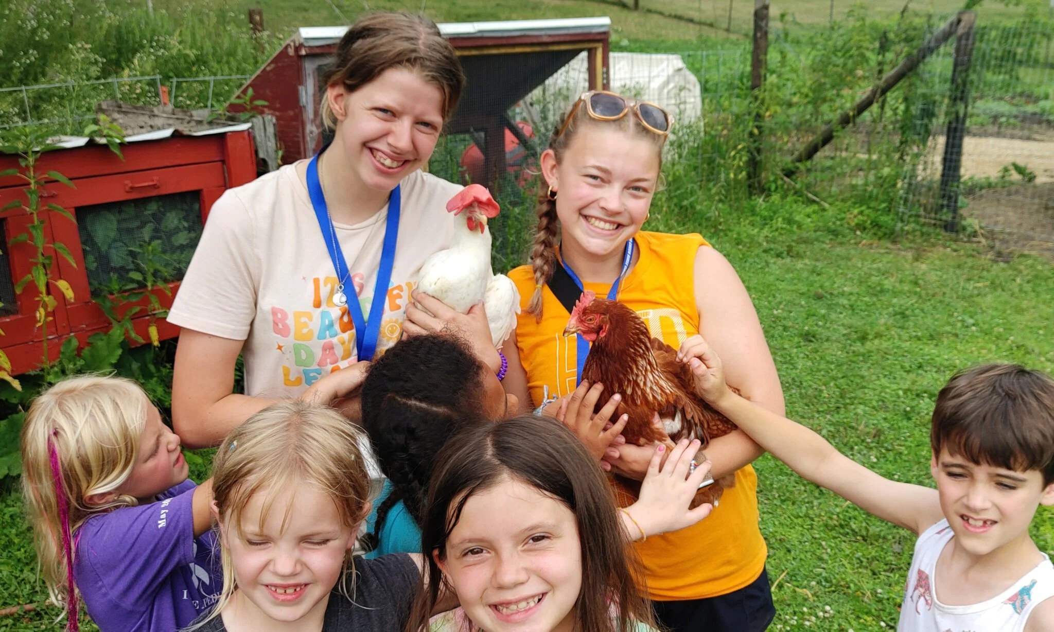 Smile of two young women holding chickens among a group of children outdoors in a garden.