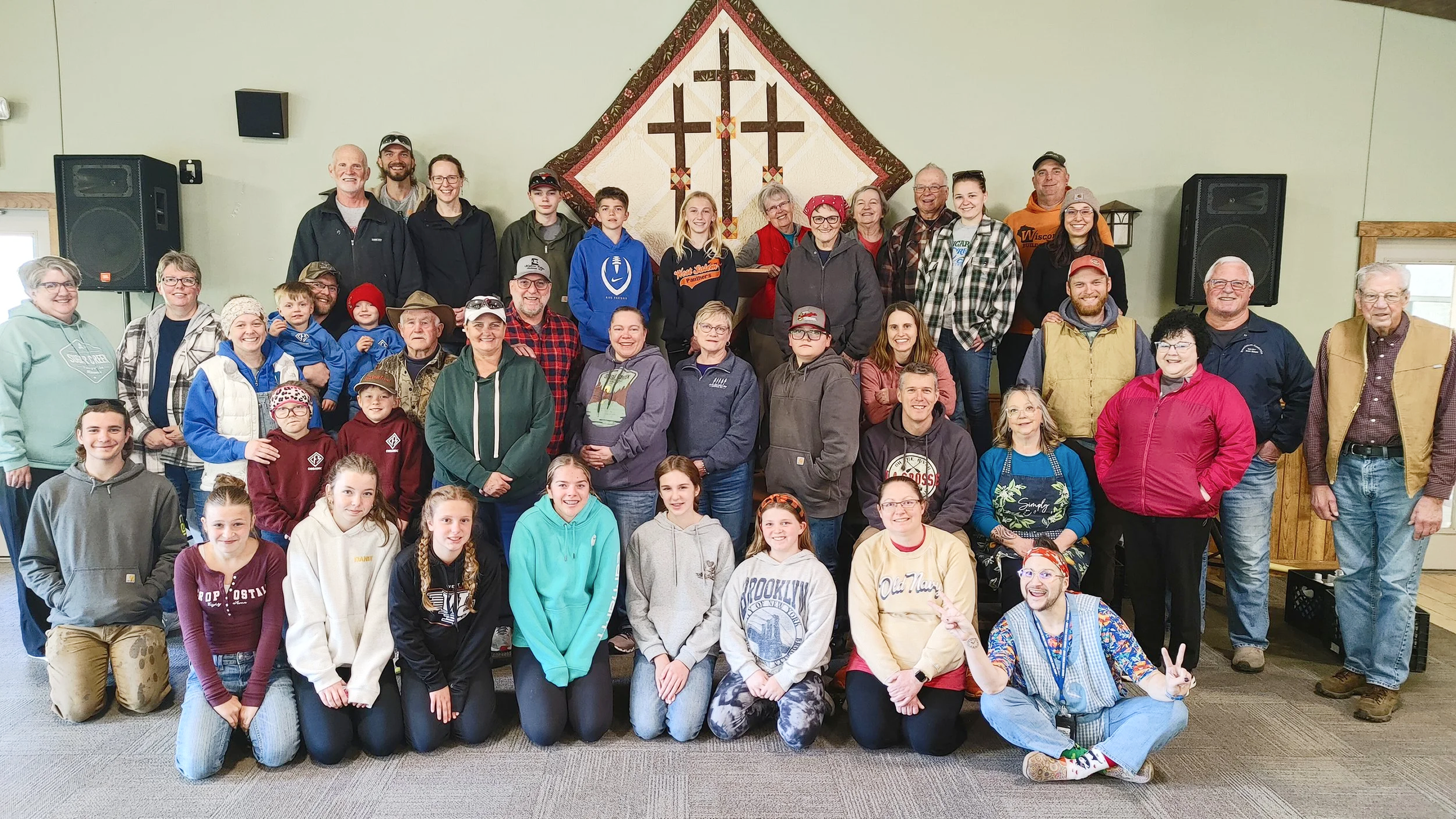 A large group of people gathered indoors for a group photo, standing in front of a wall with a decorative quilt featuring three crosses.