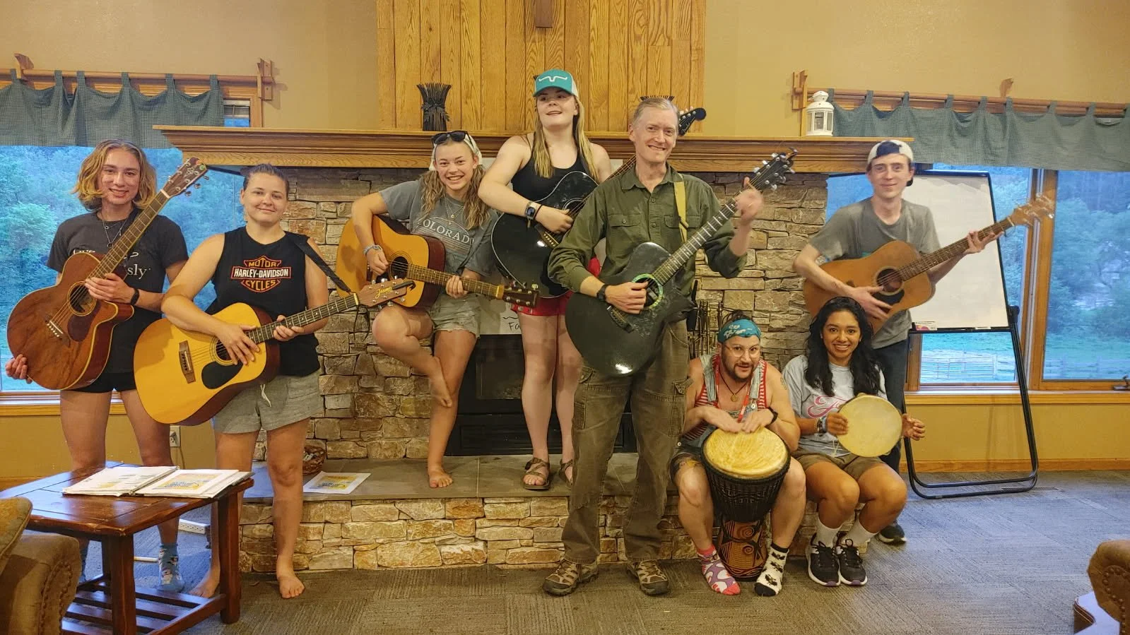 Group of nine people, eight playing guitars and one with a drum, standing and sitting in a room with a stone fireplace and large windows, smiling at the camera.