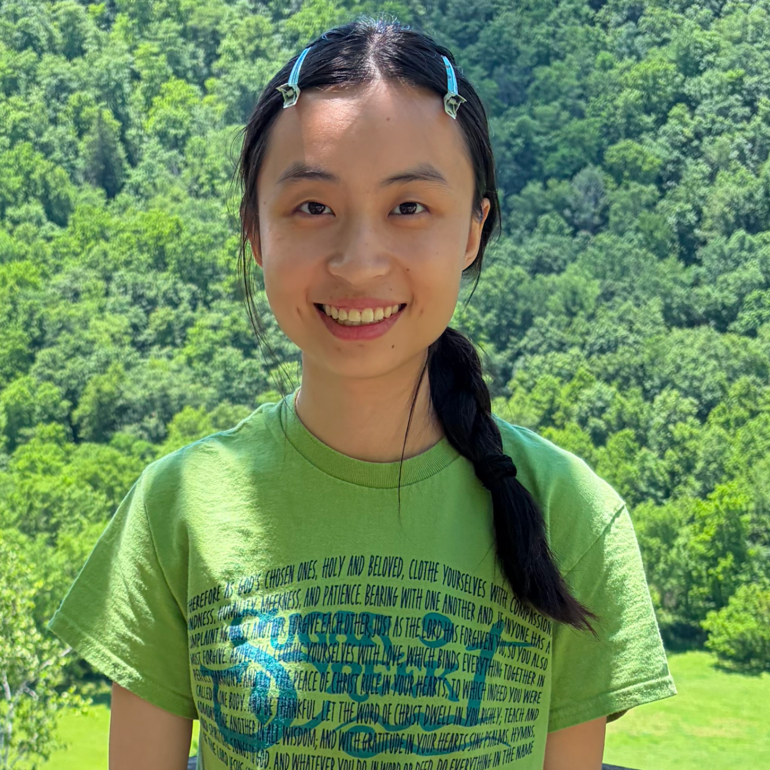 A woman outdoors in front of a lush green forest, wearing a green T-shirt with black and blue text and two blue hair clips, smiling at the camera.