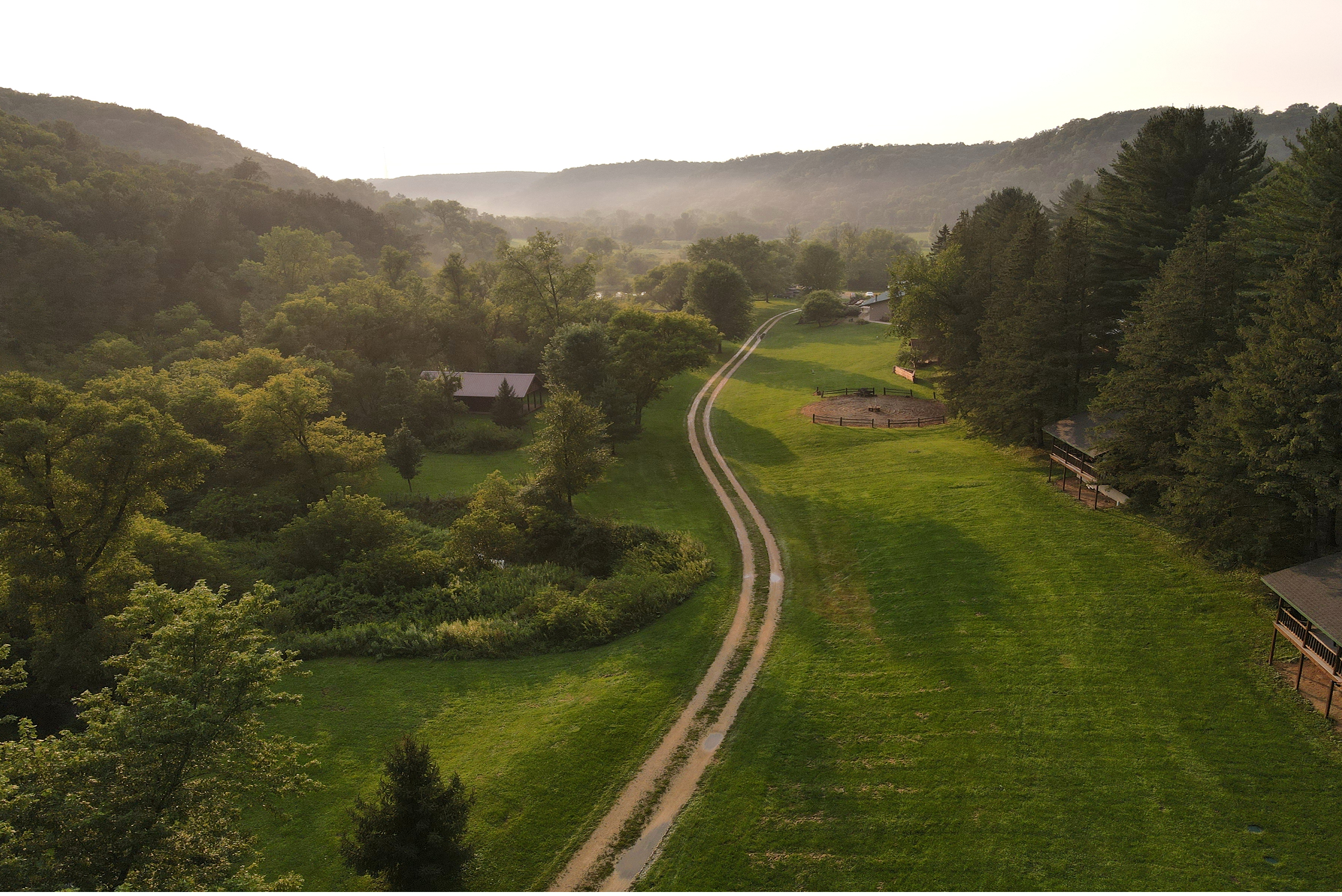 A scenic landscape of a green valley with a winding dirt road, surrounded by trees and rolling hills in the background, taken during daylight hours with clear weather.