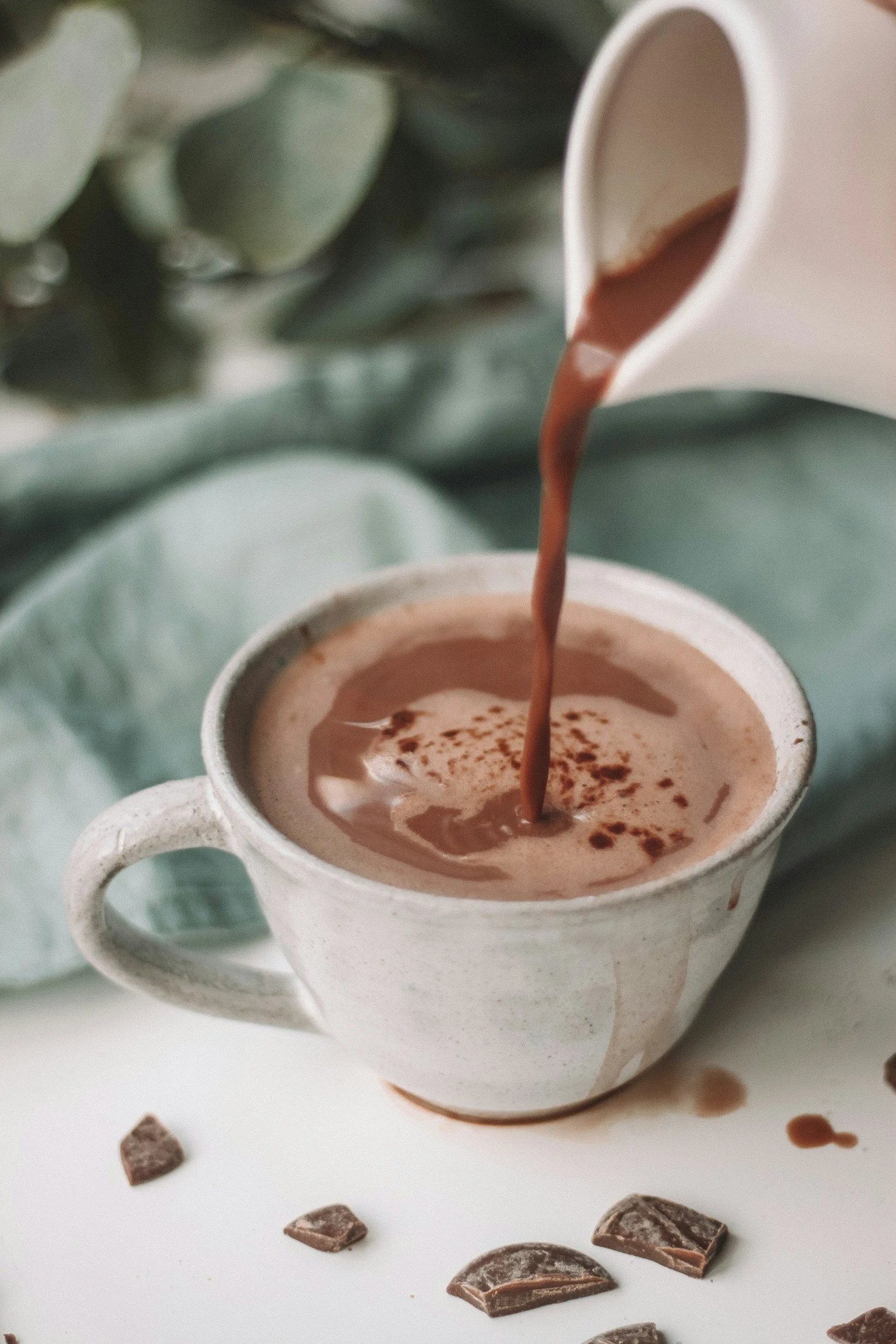 A white ceramic mug filled with hot chocolate, being poured from a white pour-over mug, with chocolate pieces scattered around on a white surface and a light-colored fabric background.
