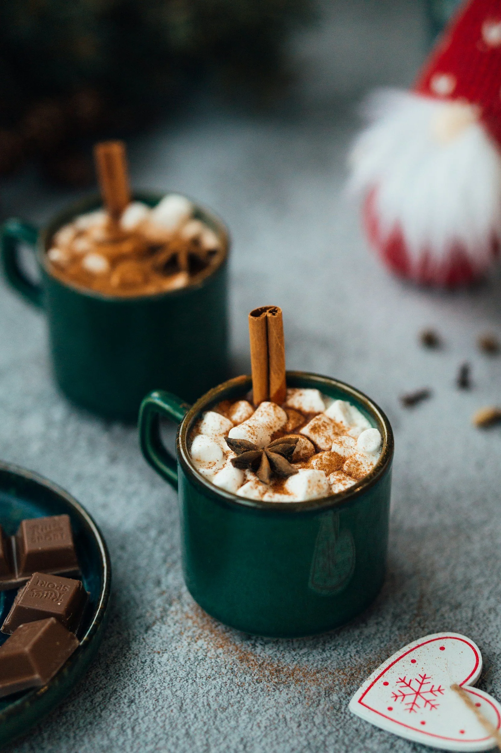 Three cups of hot chocolate topped with marshmallows and a star anise, surrounded by chocolates and festive decorations on a textured surface.