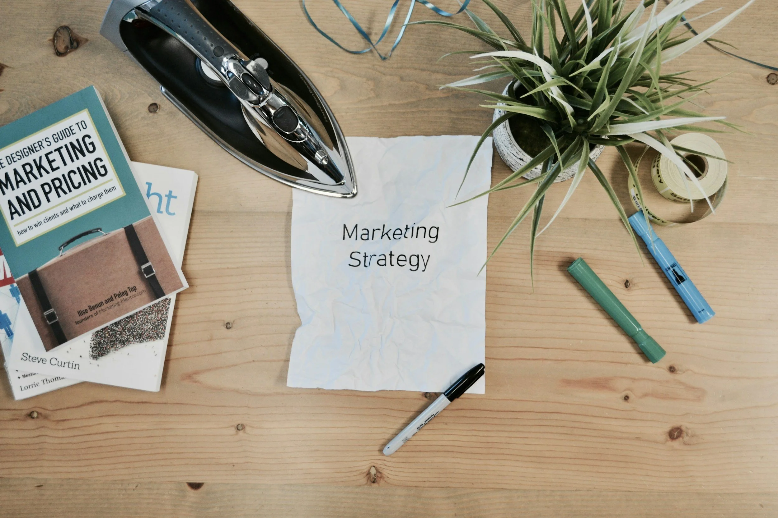 A wooden desk with an iron, a potted plant, a roll of tape, and printed papers. One paper has 'Marketing Strategy' written on it, and several pens are scattered on the desk.