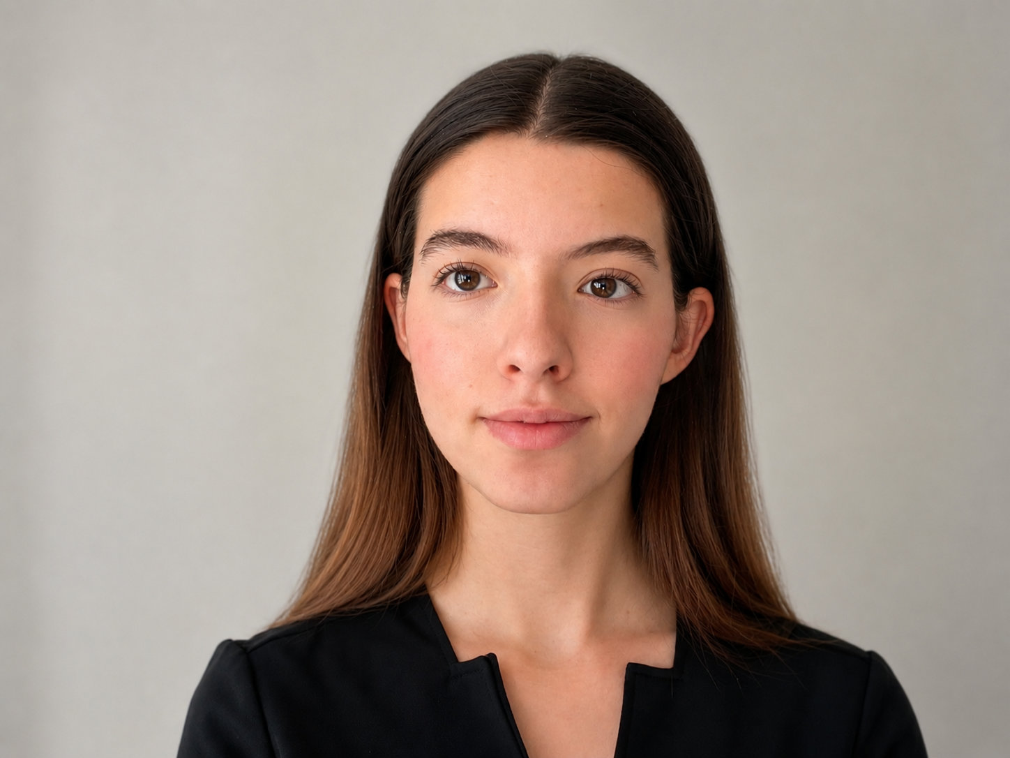 A young woman with long brown hair, wearing a black top, standing against a plain light gray background.