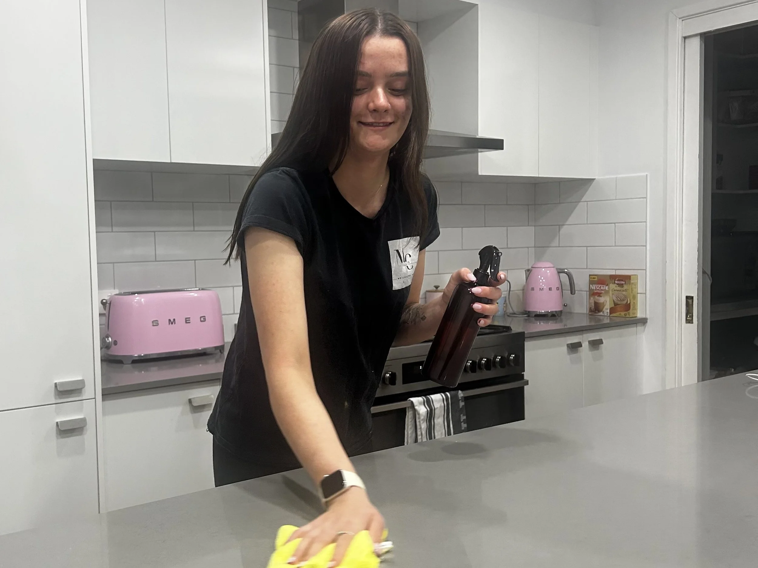 A young woman with long brown hair is smiling while cleaning a kitchen counter with a yellow cloth. She is wearing a black t-shirt and a smartwatch on her left wrist.