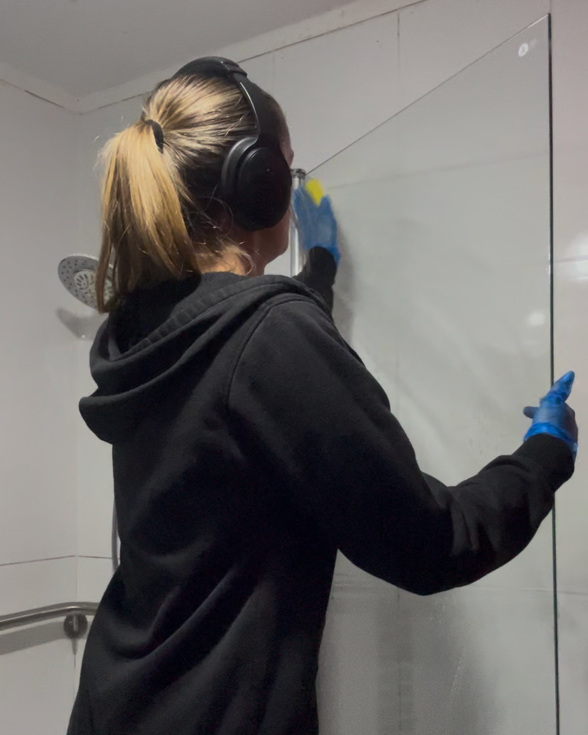 A woman wearing a black hoodie and blue gloves cleaning a glass door in a bathroom with a shower in the background.