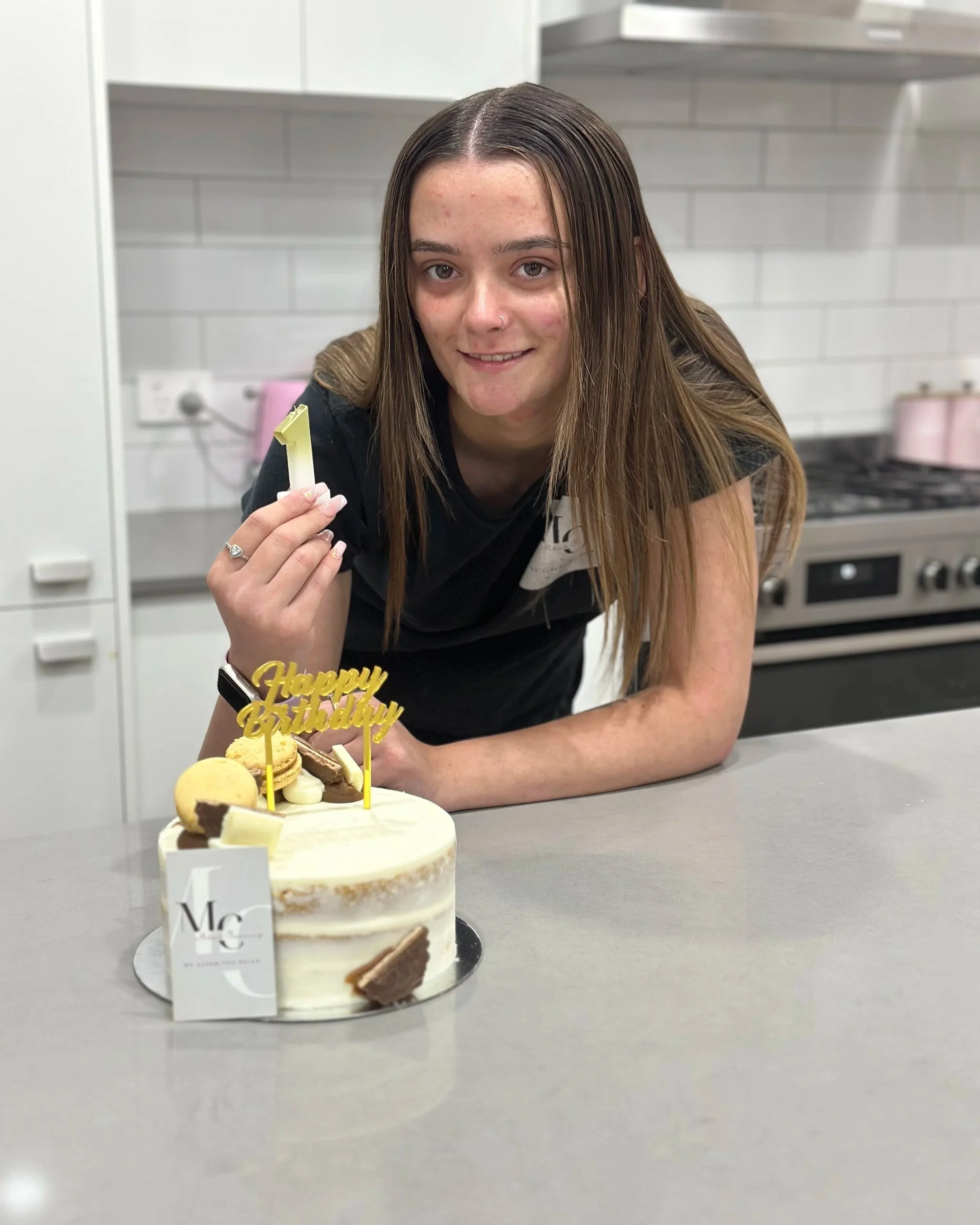 Young woman with brown hair and light skin holding a birthday candle in front of a decorated birthday cake in a kitchen.