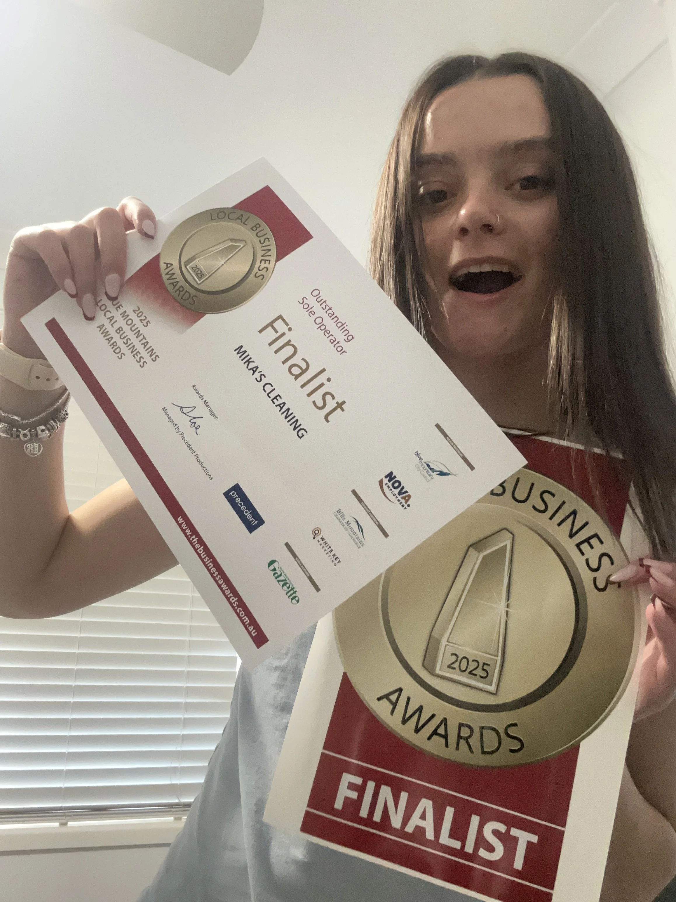 Woman holding awards and finalist certificates for local business awards with a background of blinds and a wall.