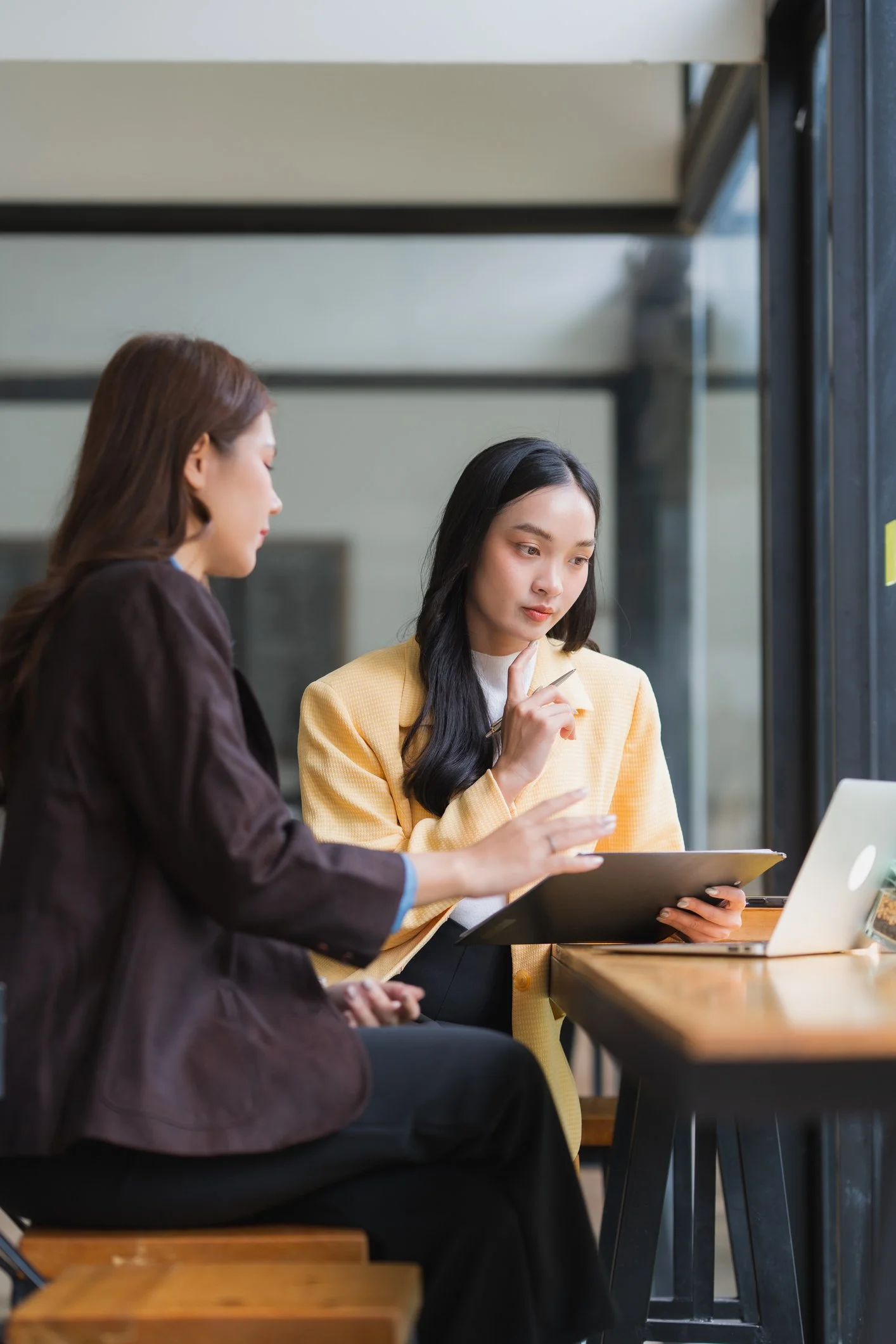 Two women sitting at a table, engaged in a discussion. One woman is wearing a brown blazer, and the other is wearing a yellow blazer, holding a folder, with a laptop nearby. They are in a modern office or cafe setting with large windows.