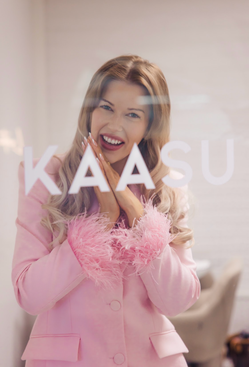 Young woman with light brown hair and pink lipstick, smiling and looking through glass with the word "KAASU" written on it, wearing a pink blazer with feathered cuffs.