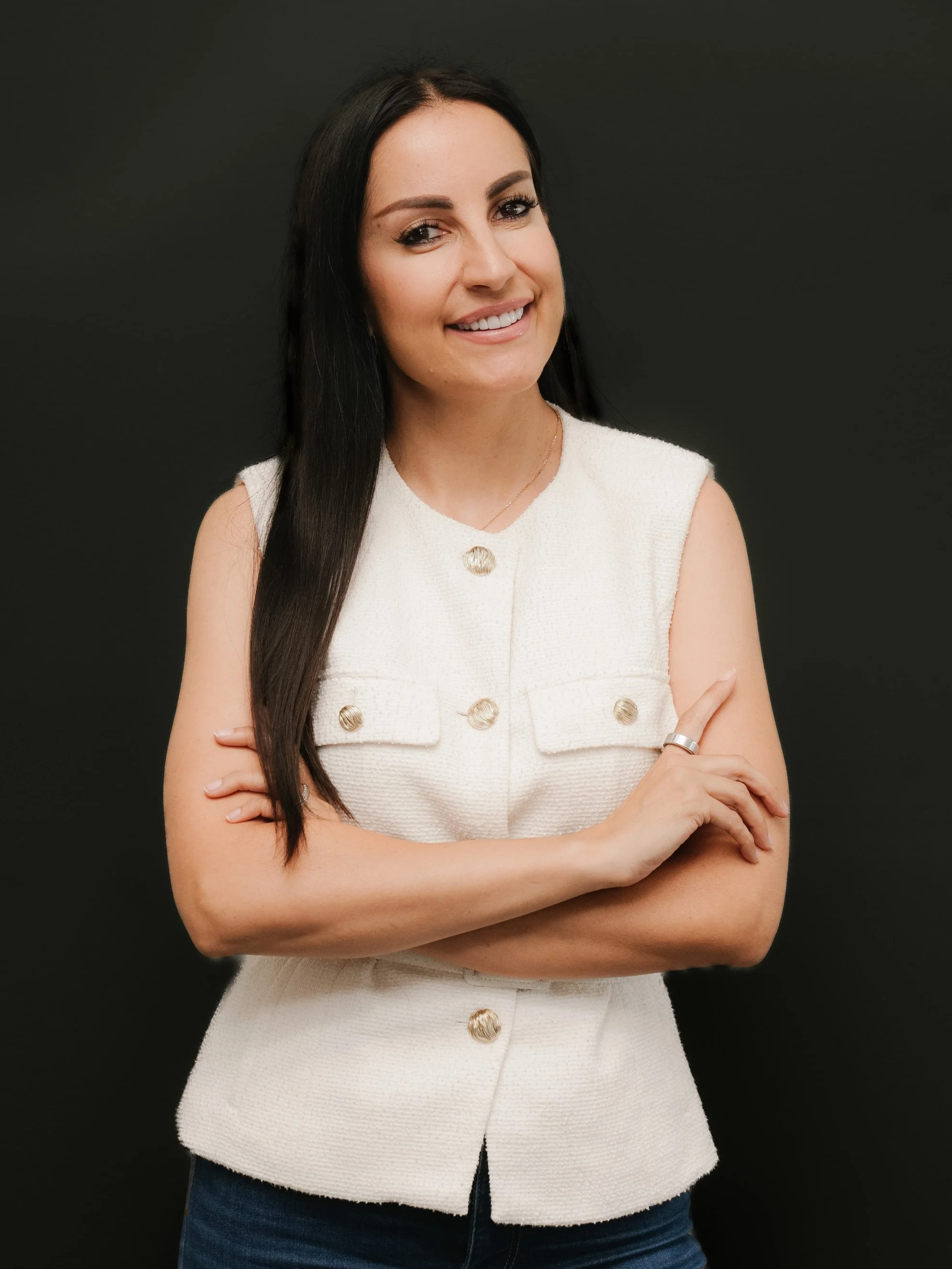 A woman with long dark hair, wearing a white sleeveless top with gold buttons, standing against a black background.