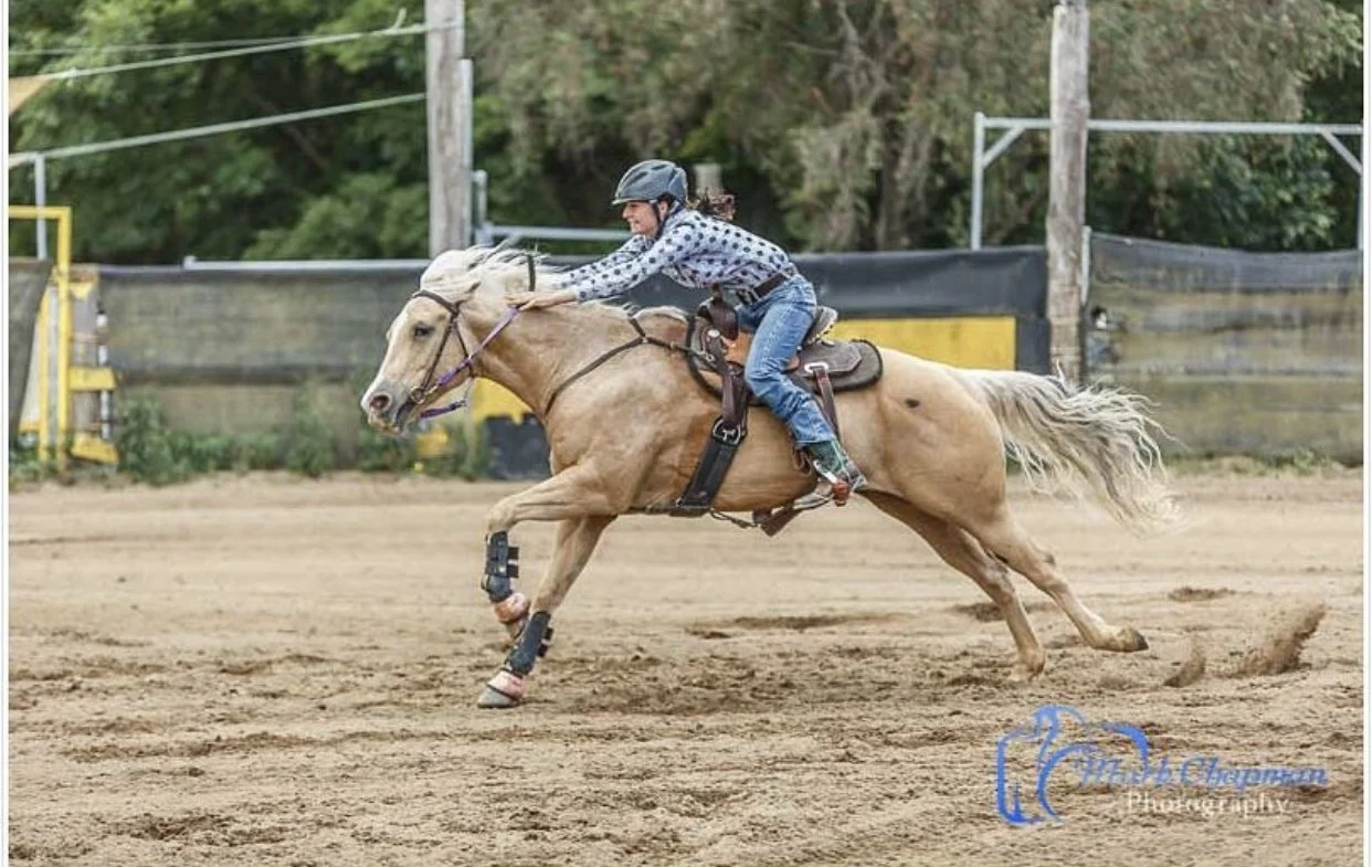 A woman riding a galloping horse on a dirt track, wearing a helmet, jeans, and a patterned long sleeve shirt, with the horse equipped with protective leg gear and a saddle.