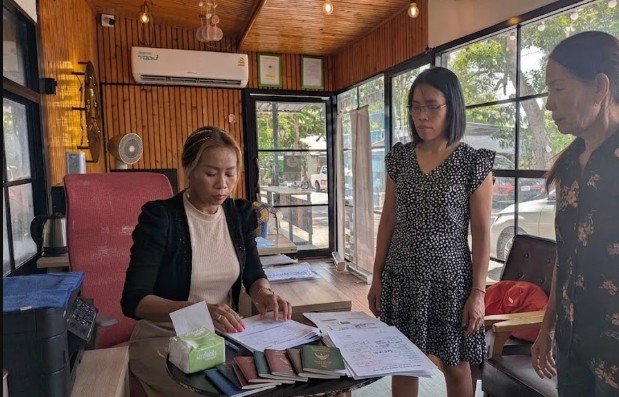 Three women in an office, one sitting at a desk and two standing, surrounded by papers and office supplies.