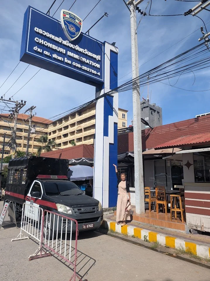 A woman standing next to a sign for the Chonburi Immigration Office, pointing upward with her right hand. There is a parked black pickup truck with a cage in the back in front of the sign. The scene takes place outdoors on a sunny day with a blue sky and a few clouds. Buildings and electrical wires are visible in the background.