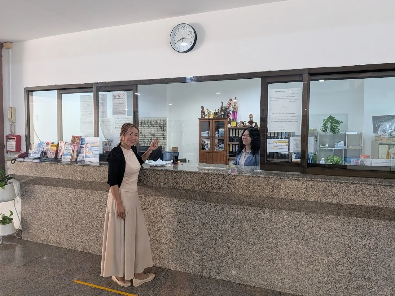 A woman in a beige dress and black cardigan smiling at a reception desk with a woman behind the glass window in an office environment.