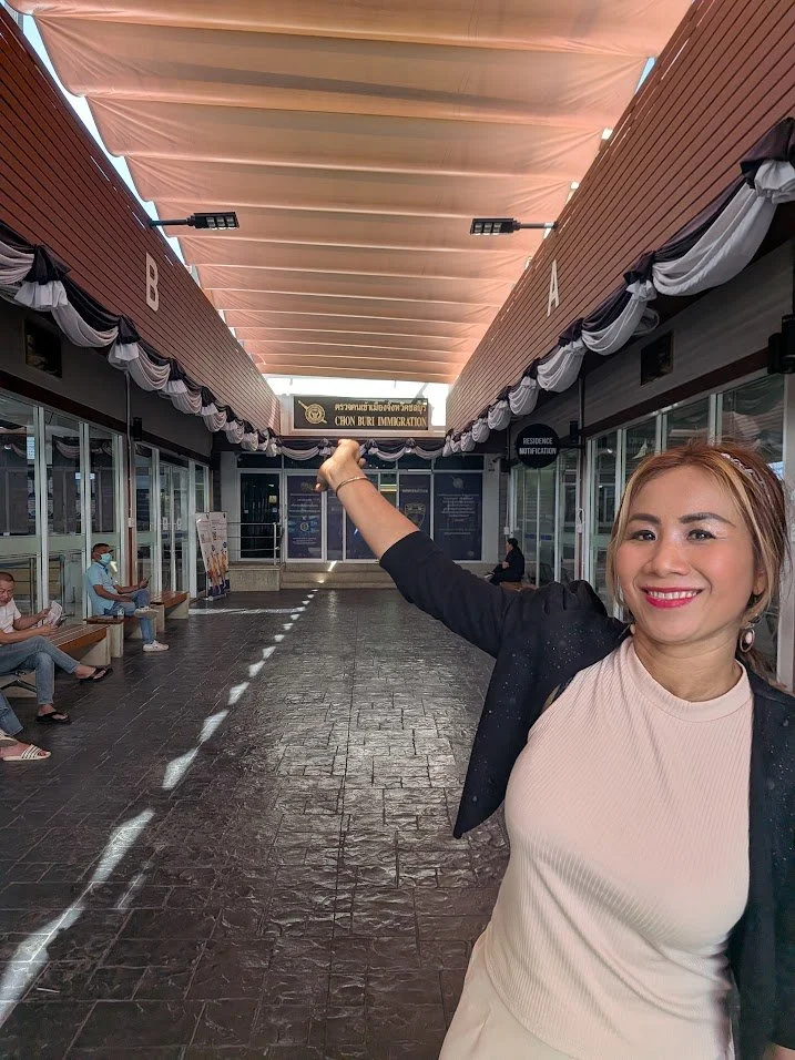 A woman smiling and pointing at the sign for 'Chon Buri Immigration' in an indoor waiting area with glass walls and benches, some people seated in the background.