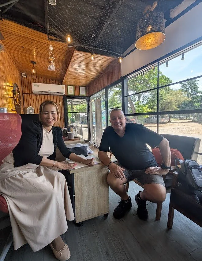 Two people sitting inside a cozy cafe with wood-paneled walls and large windows showing a sunny outdoor view, smiling at the camera.