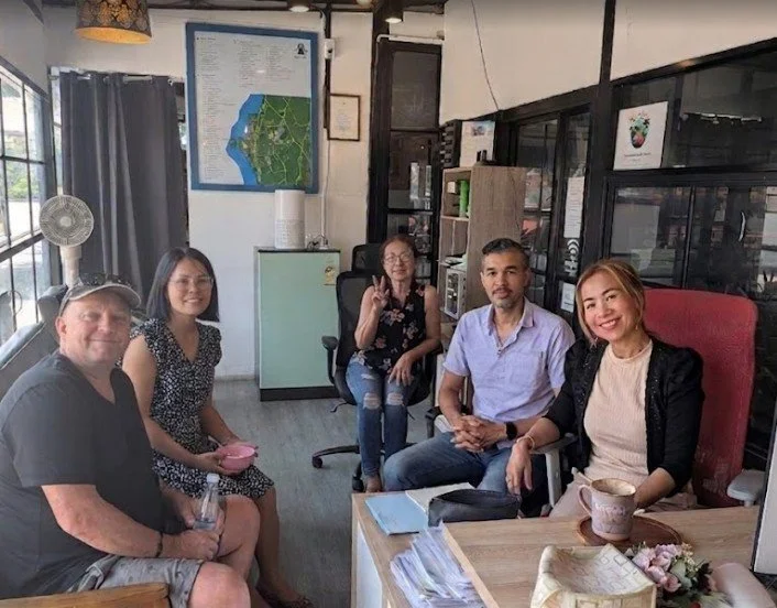 Five people sitting and smiling in an office or meeting room with shelves, a framed map on the wall, and a large window.