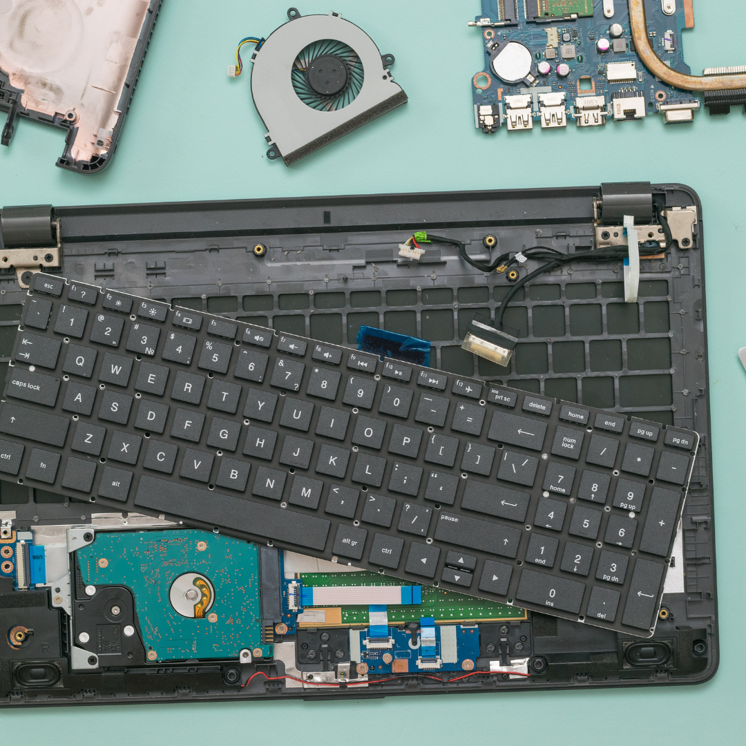 Disassembled laptop with keyboard, hard drive, circuit board, cooling fan, and other internal components spread on a light blue surface.