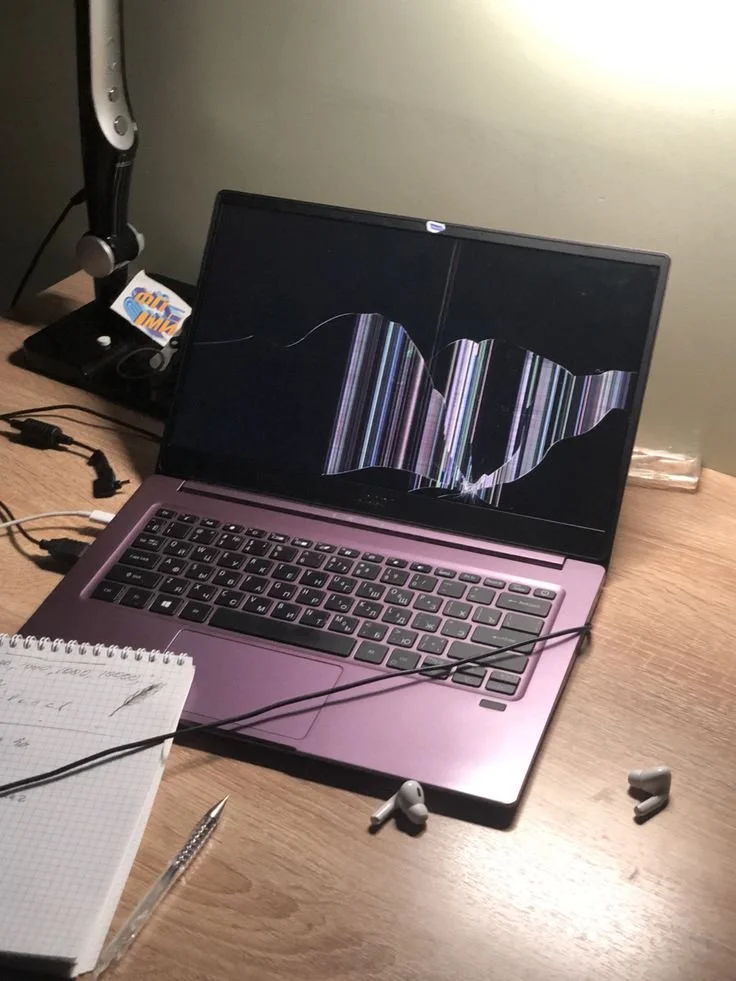 A laptop on a wooden desk with a cracked screen, white earbuds, a notebook, and a pen. There is a desk lamp and a black item with a sticky note behind the laptop.