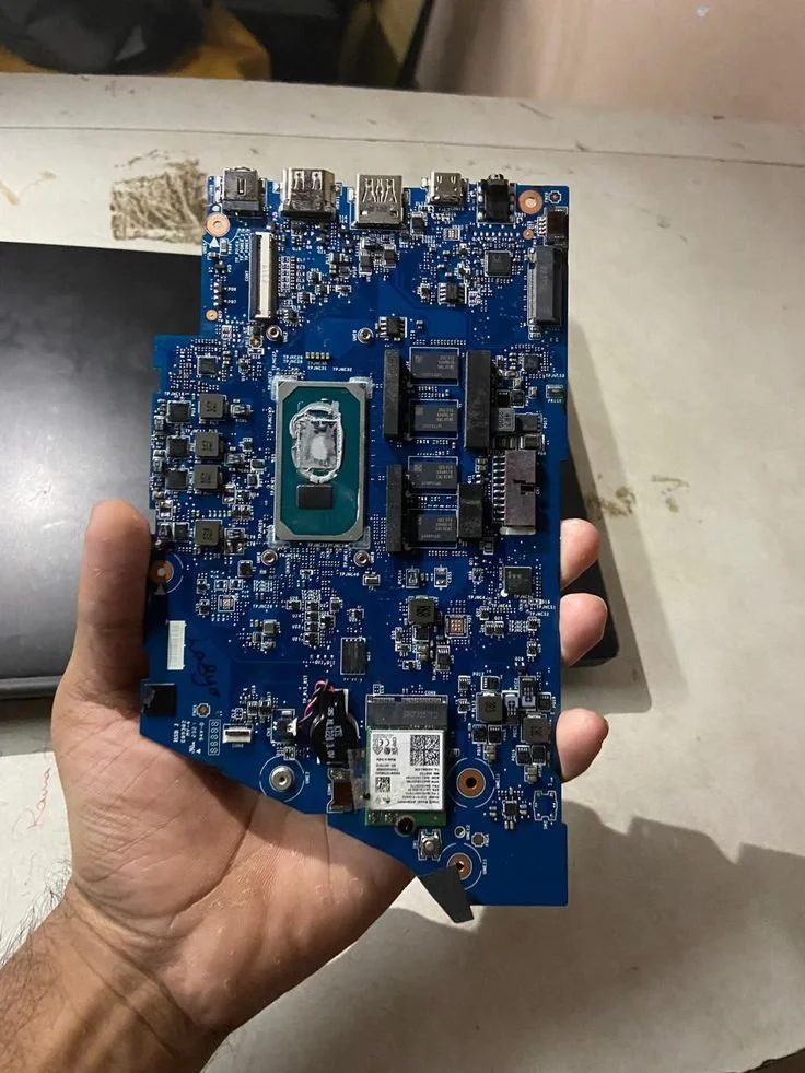 A person holding a blue computer motherboard with various electronic components and connectors, including a CPU socket, capacitors, and chips.