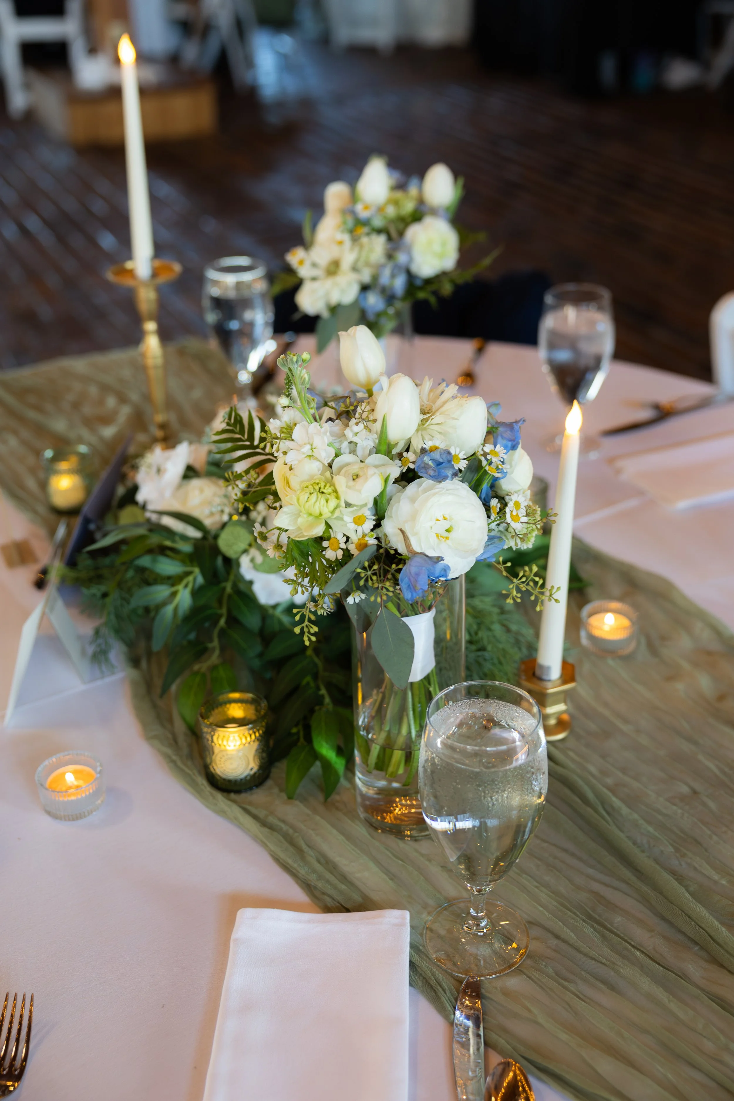A table set for a dinner party with a central flower arrangement, tall candles, water glasses, and cutlery, placed on a wooden table with a white tablecloth and a green runner.