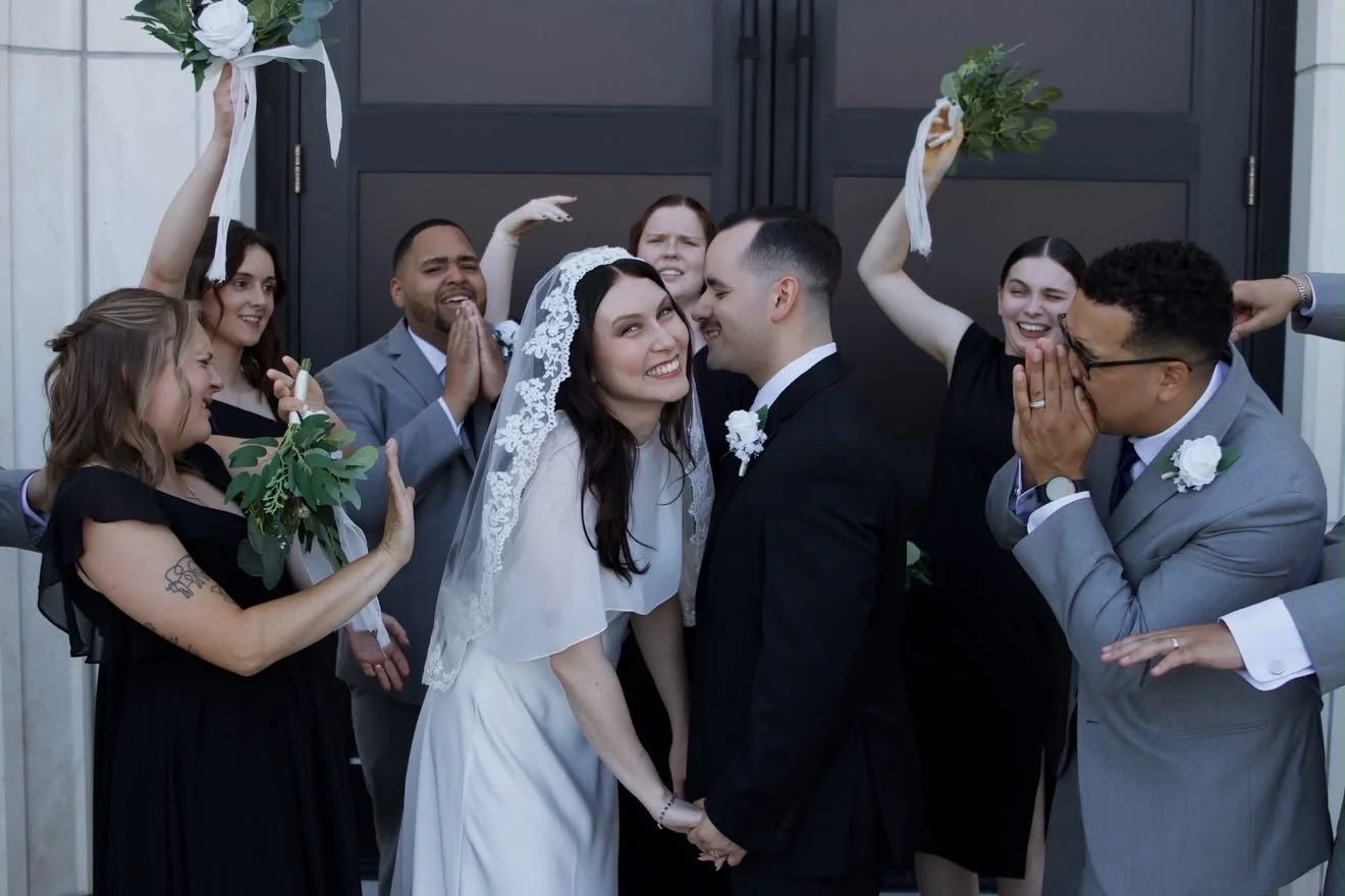 A wedding couple holding hands and smiling in front of a black door, surrounded by bridesmaids and groomsmen celebrating.